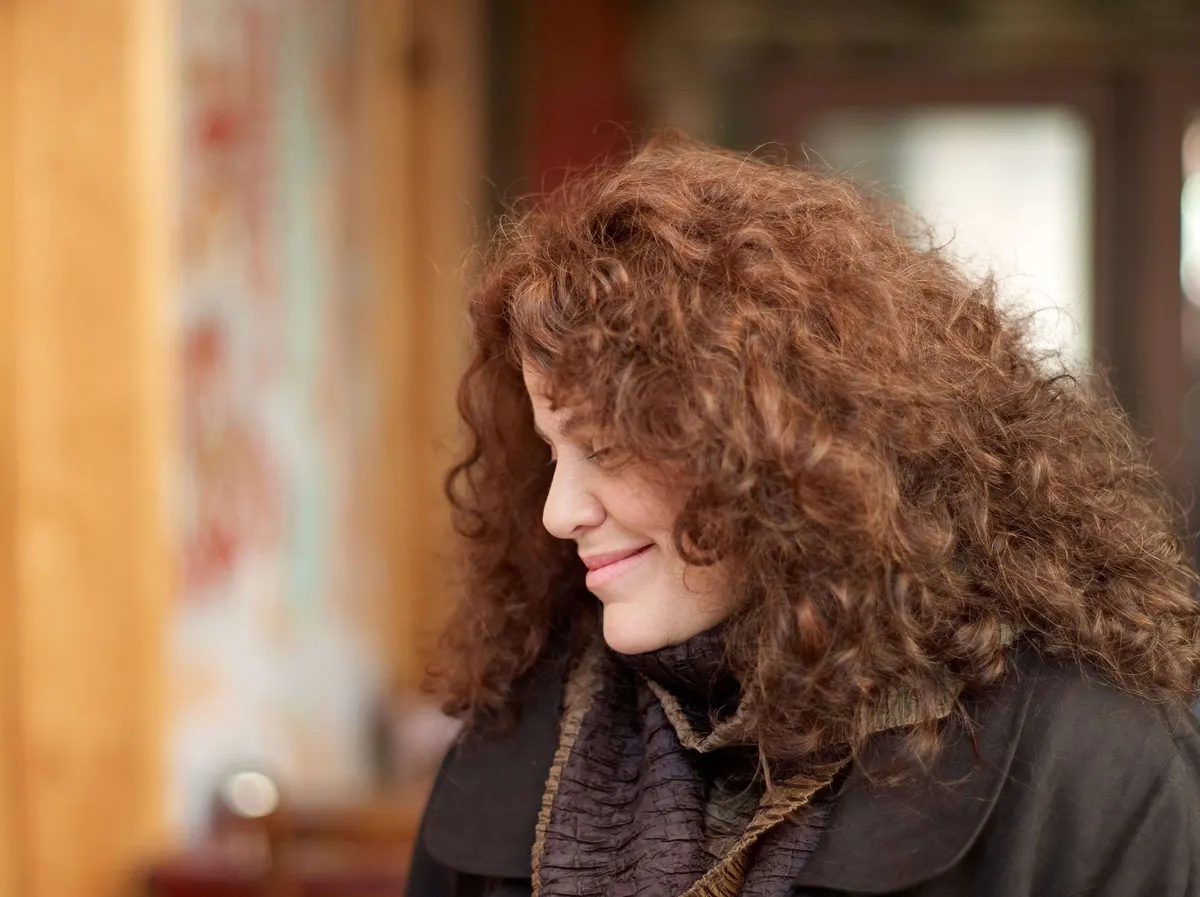 Person with curly hair wearing layered dark clothing, standing indoors with wooden furniture and soft lighting.