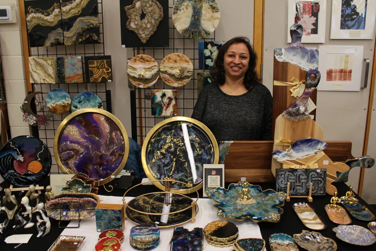 Smiling artist behind a display of colourful handmade resin trays, sculptures, and wall art at an indoor craft sale.
